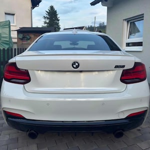 K&ouml;nnte beinhalten: Die R&uuml;ckansicht einer wei&szlig;en BMW Limousine mit einem schwarzen B58-Emblem auf dem Kofferraumdeckel, einem zentralen BMW-Logo, dunkelroten R&uuml;ckleuchten und einem schwarzen unteren Sto&szlig;f&auml;ngerdiffusor mit zwei runden Auspuffrohren.