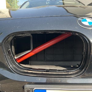 May include: Close-up of a black BMW car's front end, showing the grille and a bright red support bar. The BMW emblem is visible. The car's bonnet is open, revealing the engine compartment.