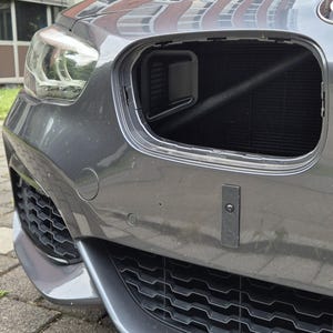 Pode incluir: Close-up de um para-choque cinza com uma grelha preta e uma seção aberta revelando o radiador. O farol do carro é visível à esquerda. O para-choque tem um design moderno.