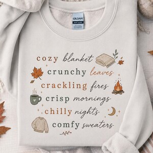 May include: A light beige crewneck sweatshirt featuring a graphic print with the text: cozy blanket, crunchy leaves, crackling fires, crisp mornings, chilly nights, comfy sweaters, accompanied by illustrations of a blanket, leaves, fire, mug, and sweater.