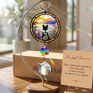 May include: A round stained glass suncatcher featuring a black cat silhouette with angel wings, the name "Bob", and a colorful landscape. It hangs from a metal stand with a crystal prism and rainbow-colored accents. A card reads "The Light Remains".