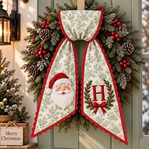 Peut inclure: Une couronne de Noël festive avec des aiguilles de pin vertes, des pommes de pin et des baies rouges. Une bannière en tissu à motif floral vert et bords festonnés rouges est suspendue au centre, avec un visage de Père Noël et un H rouge.