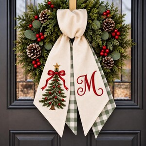 May include: A festive wreath composed of green cedar foliage, pinecones, and red berries, adorned with a cream fabric bow. One tail of the bow features a printed Christmas tree with a red ribbon and gold star, while the other displays a red letter M.