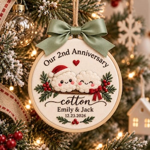 May include: A round embroidery hoop ornament featuring the text Our 2nd Anniversary, cotton, Emily & Jack, and 12.23.2026. It displays two white fluffy characters, one wearing a red Santa hat, with green holly, red berries, and a sage green bow.