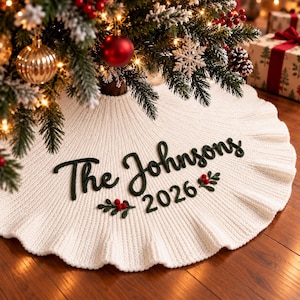 Pode incluir: Uma saia de árvore de Natal em malha branca com borda franzida, apresentando texto bordado em verde escuro que diz The Johnsons e o ano 2026, acentuado com pequenos detalhes bordados de bagas vermelhas e folhas verdes sob o texto.