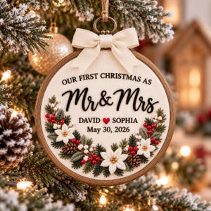 Puede incluir: Un adorno navideño en bastidor de madera con tela color crema que muestra el texto negro "OUR FIRST CHRISTMAS AS Mr & Mrs", "DAVID" y "SOPHIA" separados por un corazón rojo, y la fecha "May 30, 2026". Decorado con flores de pascua, piñas y bayas rojas bordadas, rematado con un lazo de tela color crema.