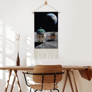 May include: A wall hanging featuring a whimsical cartoon character wearing a hat, standing on the moon with the Earth in the background. The image includes a lunar lander and an American flag. The bottom of the hanging has a cream-colored fringe.