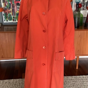 May include: A bright orange long-sleeve coat featuring a wide collar, a front button closure with five matching orange buttons, and two rectangular front pockets. The garment is accented with decorative white topstitching along the collar, pockets, and seams.