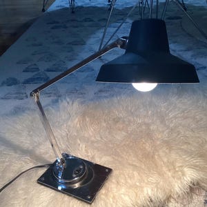 May include: A black desk lamp with a chrome-colored adjustable arm and base. The lamp has a cone-shaped shade and a bright lightbulb. The base is square and has a power switch. The lamp is on a fluffy, white surface.