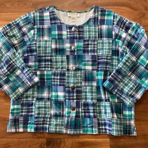 May include: A long-sleeve jacket featuring a patchwork design of various plaid patterns in shades of blue, teal, green, and white. The jacket has a round neckline, a front button closure with five metallic buttons, and an Orvis brand label inside.
