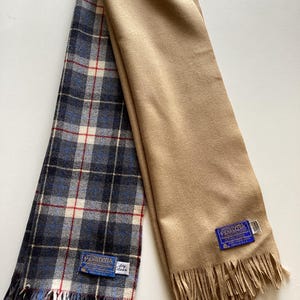 May include: A reversible scarf featuring a solid tan side and a plaid side with blue, black, white, and red lines. Both ends have fringe. A blue Pendleton label is sewn onto each side, with a small white tag reading dry clean on the plaid side.