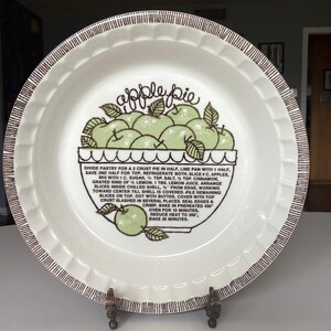 May include: White ceramic pie plate with a brown rim and a decorative stand. The center features a green apple illustration with the words "apple pie" and a recipe. The plate is designed for baking and serving homemade pies.