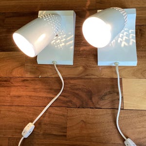 May include: A pair of white wall-mounted lamps featuring cylindrical heads with perforated detailing. Each lamp has a rectangular base, a white power cord with an inline switch, and emits a bright light, casting patterned shadows on the surface.