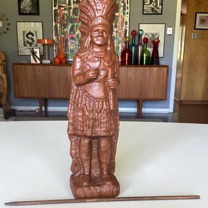 May include: A brown carved wooden figurine depicting a person wearing a feathered headdress and traditional attire, standing on a rectangular base. A thin, straight wooden rod is positioned horizontally on the surface in front of the figurine.