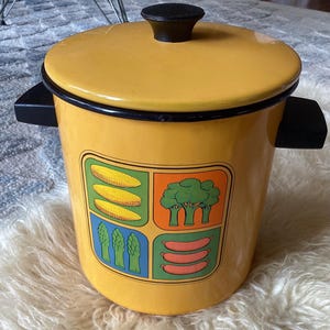 May include: Yellow enamel pot with a lid and black handles. The pot features a colorful graphic of vegetables, including corn, broccoli, asparagus, and sausages. The lid has a black knob.