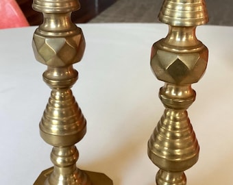 Könnte beinhalten: Zwei antike Messingkerzenhalter. Jeder Kerzenhalter hat einen quadratischen Sockel, einen detaillierten Schaft mit geometrischen und abgerundeten Formen und eine kreisförmige Oberseite für eine Kerze. Die Kerzenhalter sind aus glänzendem, goldenem Messing.