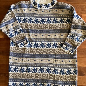 May include: A long-sleeved knit sweater featuring a repeating horizontal pattern in shades of cream, navy blue, and tan. The design includes bands of stylized leaves, wavy lines, and small heart shapes, finished with a ribbed mock neck collar.
