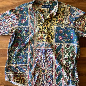 May include: A short-sleeve Ralph Lauren polo shirt featuring a multi-colored patchwork pattern with floral and paisley motifs in shades of blue, yellow, red, and green. The shirt has a button-down collar and a chest pocket on the right side.