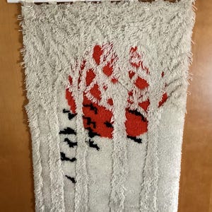 May include: A rectangular wall hanging made of shaggy, off-white yarn featuring a central design of a large, textured red circle partially obscured by vertical off-white strips and small black markings that resemble tree trunks and branches.