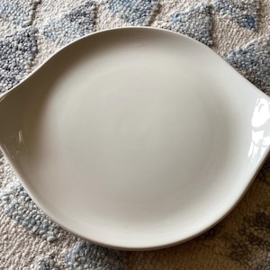 May include: A white ceramic serving platter with a unique, curved shape. The platter has two integrated handles and a smooth, glossy finish. This dish is ideal for serving appetizers or small dishes.