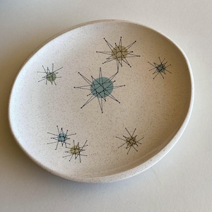 May include: Off-white ceramic plate with a speckled surface. The plate features a mid-century modern design with starburst patterns in teal, yellow, and black. The plate has an oval shape.
