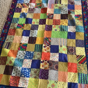 May include: A patchwork quilt featuring a grid of multicolored fabric squares in various patterns including stripes, dots, and floral prints. The quilt is bordered by a dark blue fabric with a repeating colorful geometric motif.