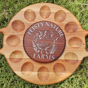 Op de afbeelding: Houten eierplateau met een cirkelvormig ontwerp, met 12 eivormige inkepingen. Een kleinere, ronde, donkere houten inzet in het midden toont de tekst "FEISTY NATURE FARMS" en een illustratie van een kip. De tray heeft twee handvatten.