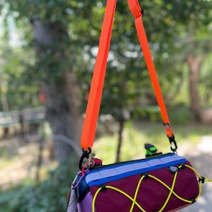 May include: A colorful cylindrical bag with an orange shoulder strap. The bag features a burgundy body with yellow bungee cords, purple and blue accents, and a small logo patch. The bag is suspended against a blurred green background.