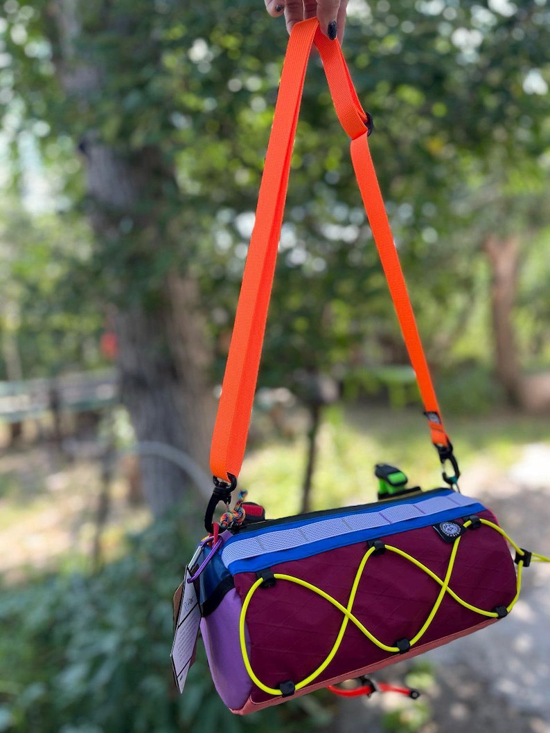 May include: A colorful cylindrical bag with an orange shoulder strap. The bag features a burgundy body with yellow bungee cords, purple and blue accents, and a small logo patch. The bag is suspended against a blurred green background.