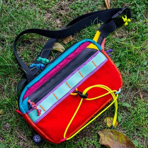 May include: A colorful fanny pack with a black strap, lying on grass. The bag features a red base, with sections of black, pink, teal, and yellow. The bag has a black zipper and yellow cord details.