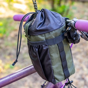 May include: A black and olive green bicycle handlebar bag with a drawstring closure. The bag is attached to purple handlebars with purple grips. The bag has a black strap and a small pocket. The background is blurred green foliage.