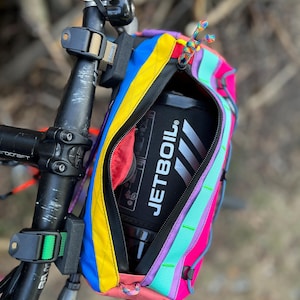 May include: A colorful bicycle handlebar bag with a zippered opening, featuring a vibrant array of colors including blue, yellow, pink, and purple. The bag is attached to the handlebars and contains a black Jetboil container. The bag has a rainbow zipper pull.