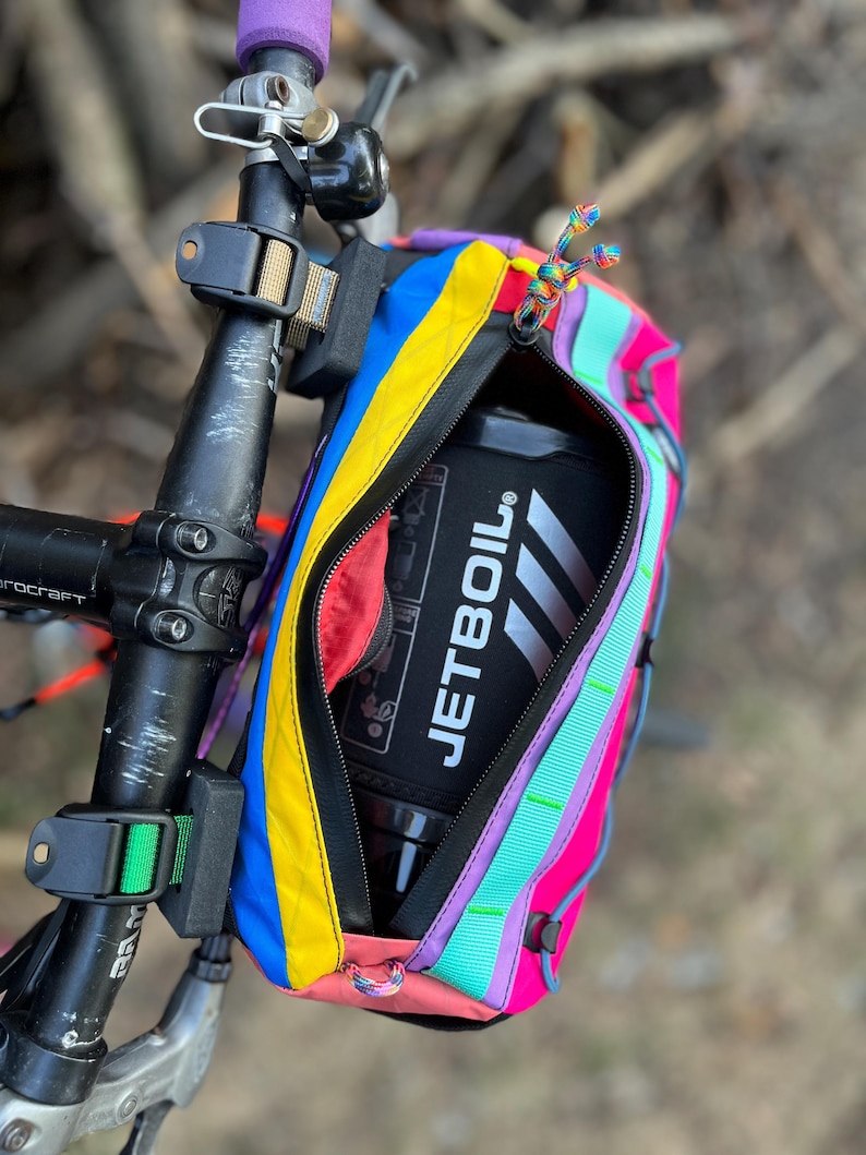 May include: A colorful bicycle handlebar bag with a zippered opening, featuring a vibrant array of colors including blue, yellow, pink, and purple. The bag is attached to the handlebars and contains a black Jetboil container. The bag has a rainbow zipper pull.