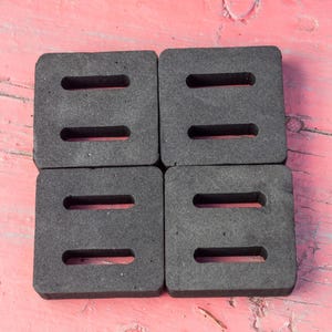 May include: Four dark gray, square foam pads with three rectangular cutouts each, arranged in a 2x2 grid. The pads are on a weathered, pink wooden surface.