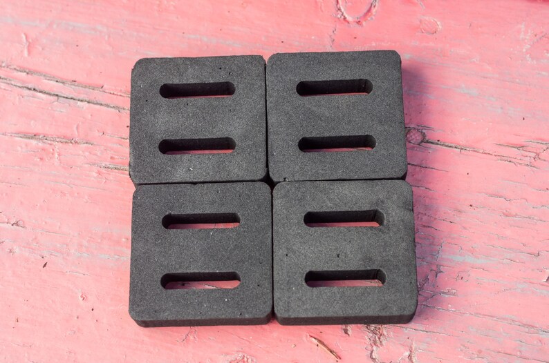 May include: Four dark gray, square foam pads with three rectangular cutouts each, arranged in a 2x2 grid. The pads are on a weathered, pink wooden surface.