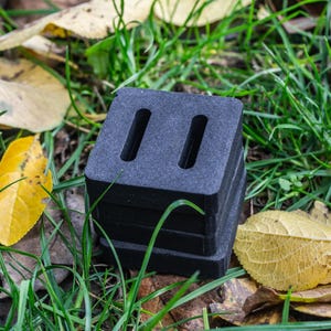 May include: A stack of four black, square-shaped blocks with two vertical slots cut into the top block. The blocks are resting on a bed of green grass and fallen yellow leaves.