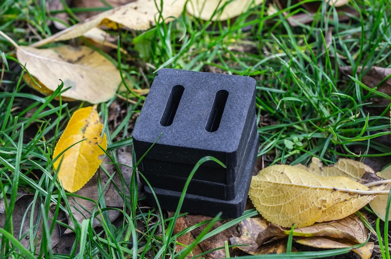 May include: A stack of four black, square-shaped blocks with two vertical slots cut into the top block. The blocks are resting on a bed of green grass and fallen yellow leaves.