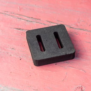 May include: A dark gray, square rubber object with two rectangular cutouts. The object is set against a weathered, pink wooden surface. The rubber piece appears to be a component or part.