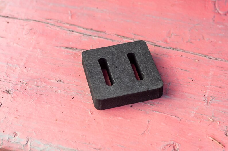 May include: A dark gray, square rubber object with two rectangular cutouts. The object is set against a weathered, pink wooden surface. The rubber piece appears to be a component or part.