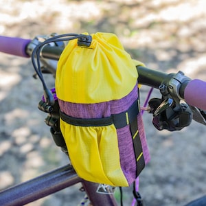Puede incluir: Una bolsa de manillar de bicicleta amarilla y morada está sujeta al manillar de una bicicleta morada. La bolsa tiene un cierre de cordón y una correa negra. El manillar tiene empuñaduras moradas.
