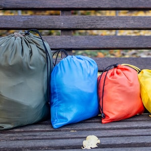 Peut inclure: Quatre sacs à cordon colorés, vert olive, bleu, rouge et jaune, avec des cordons noirs, sont posés sur un banc en bois vieilli. Les sacs sont faits d'un matériau texturé et léger, idéal pour le voyage ou le stockage.