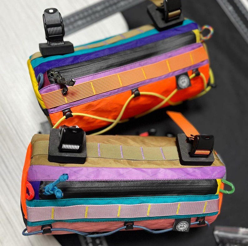 May include: Two colorful handlebar bags with a variety of colors including orange, purple, and teal. Each bag has a black zipper and black plastic clips. The bags are designed for bicycles.