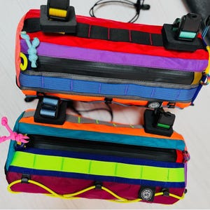 May include: Two colorful bicycle handlebar bags. The bags feature a rainbow of colors including red, purple, orange, and teal. Each bag has a black zipper and black straps with buckles. The bags are designed for attaching to a bicycle.