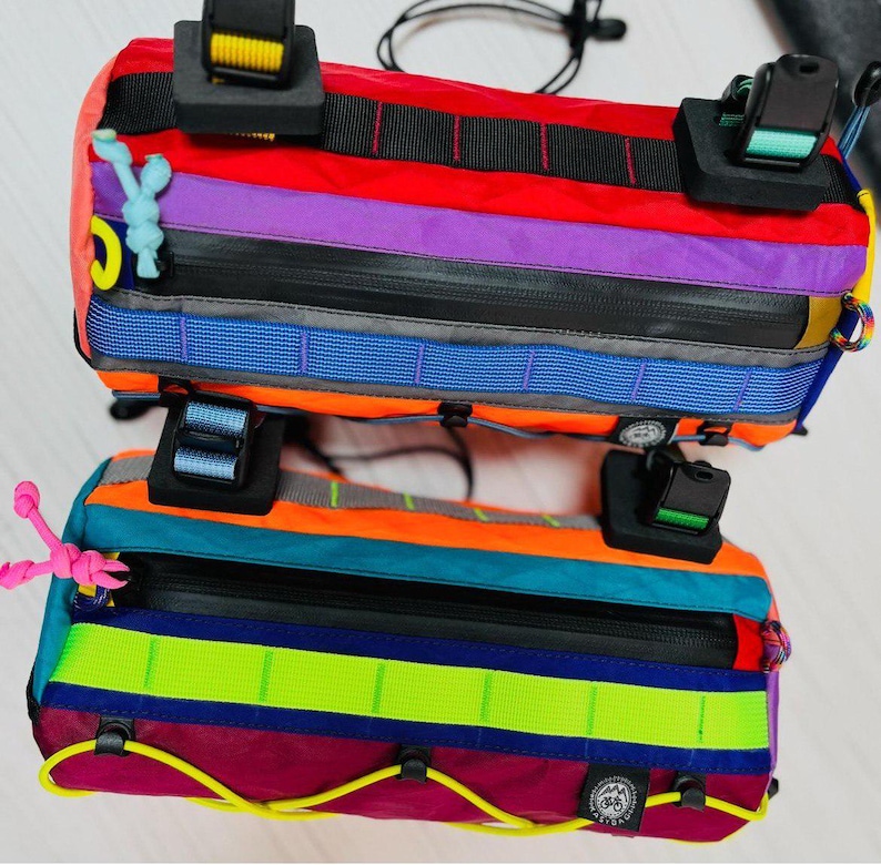 May include: Two colorful bicycle handlebar bags. The bags feature a rainbow of colors including red, purple, orange, and teal. Each bag has a black zipper and black straps with buckles. The bags are designed for attaching to a bicycle.
