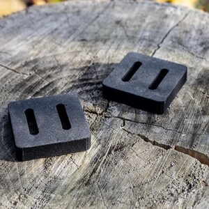 May include: Two square, black rubber pads with two rectangular cutouts each, resting on a weathered, gray tree stump. The pads are evenly spaced and appear to be new.