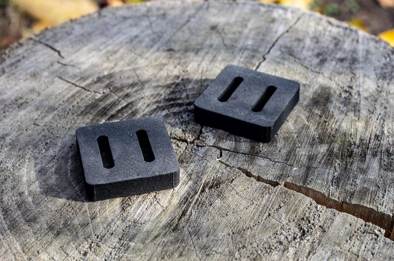 May include: Two square, black rubber pads with two rectangular cutouts each, resting on a weathered, gray tree stump. The pads are evenly spaced and appear to be new.