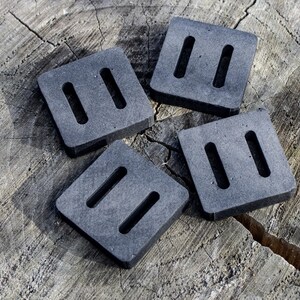 May include: Four square, dark gray weights with two rectangular slots each, arranged on a weathered, light brown wooden surface. The weights appear to be made of a dense material, possibly rubber or a similar compound. The image is well-lit, highlighting the texture of the wood.