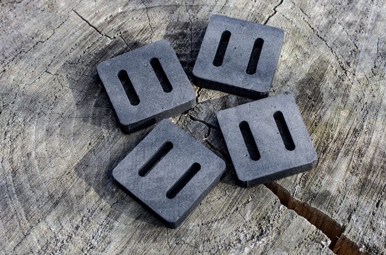 May include: Four square, dark gray weights with two rectangular slots each, arranged on a weathered, light brown wooden surface. The weights appear to be made of a dense material, possibly rubber or a similar compound. The image is well-lit, highlighting the texture of the wood.
