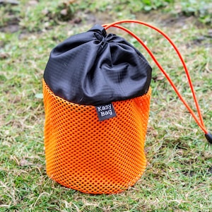 May include: An orange mesh bag with a black top and drawstring closure. The bag has a small black tag that reads "Kasy Bag". An orange drawstring extends from the top of the bag.