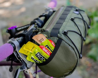 Pode incluir: Uma bolsa verde para guidão de bicicleta com alças pretas e cordas elásticas, montada em uma bicicleta. A bolsa contém embalagens de gel energético em amarelo, vermelho e bege. As manoplas do guidão são roxas.